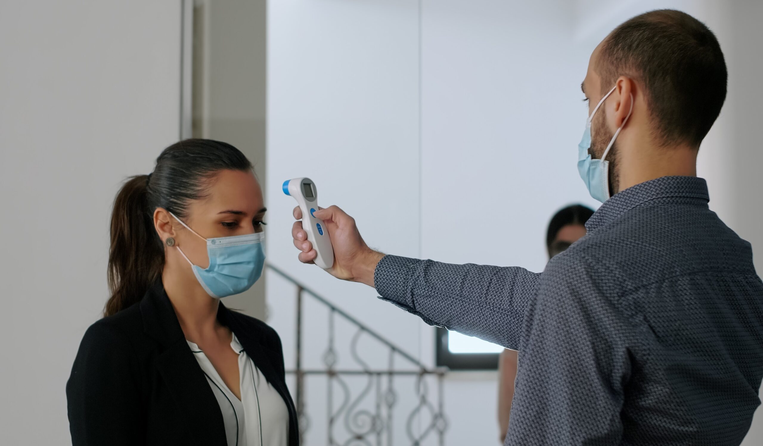 Freelancer with protection face mask measure temperature with thermometer before coworkers enter in business office. Collegues respecting social distance to avoid infection with covid19