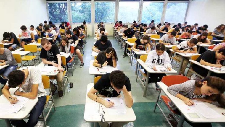 Alunos estão em uma sala com carteiras brancas realizando a prova do Exame Nacional do Ensino Médio. Todos estão de cabeça baixa.