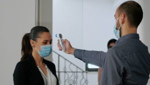 Freelancer with protection face mask measure temperature with thermometer before coworkers enter in business office. Collegues respecting social distance to avoid infection with covid19