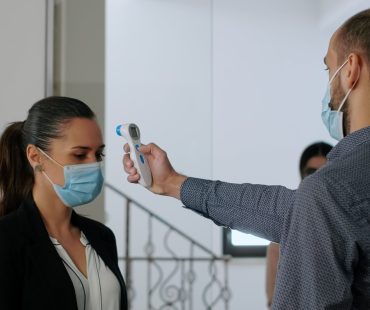 Freelancer with protection face mask measure temperature with thermometer before coworkers enter in business office. Collegues respecting social distance to avoid infection with covid19